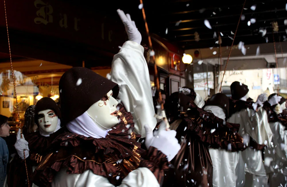 A masked Carnival band in ivory and white Pierrot costumes with copper-brown ruffs and dark hats beneath the Limoux arcades as confetti falls.